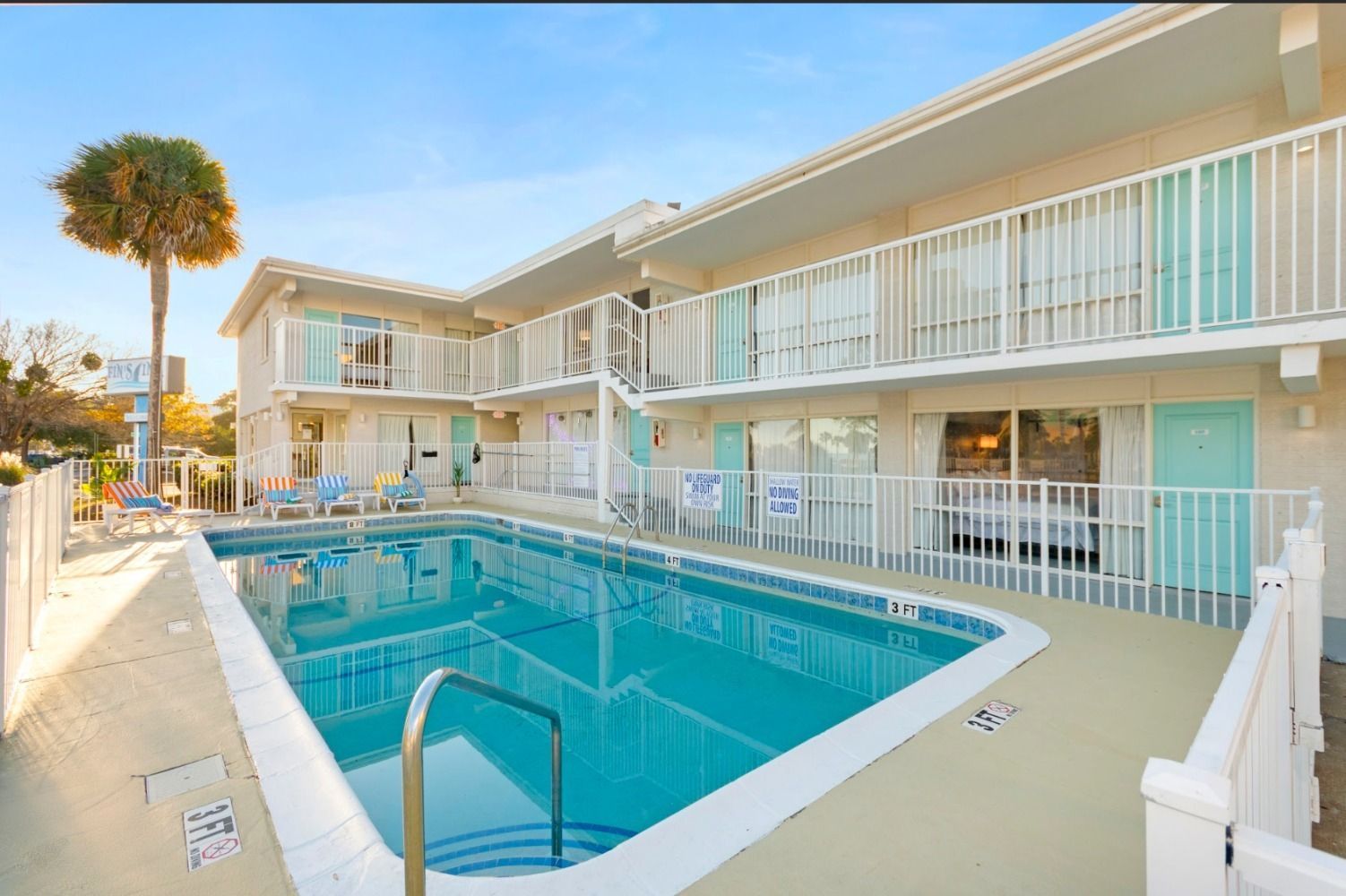 Motel with pool; two-story building, white with turquoise shutters. Palm tree, chairs, and clear blue water. Sunny day.