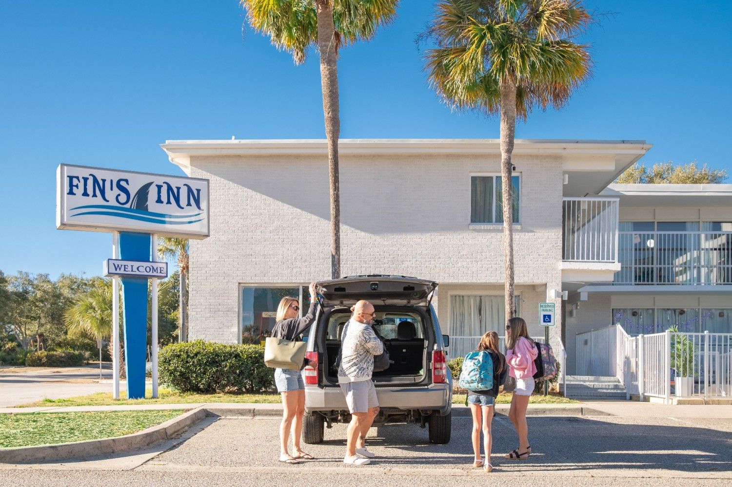 Family unloading luggage from a car in front of "Fin's Inn" motel under palm trees, bright sunny day.
