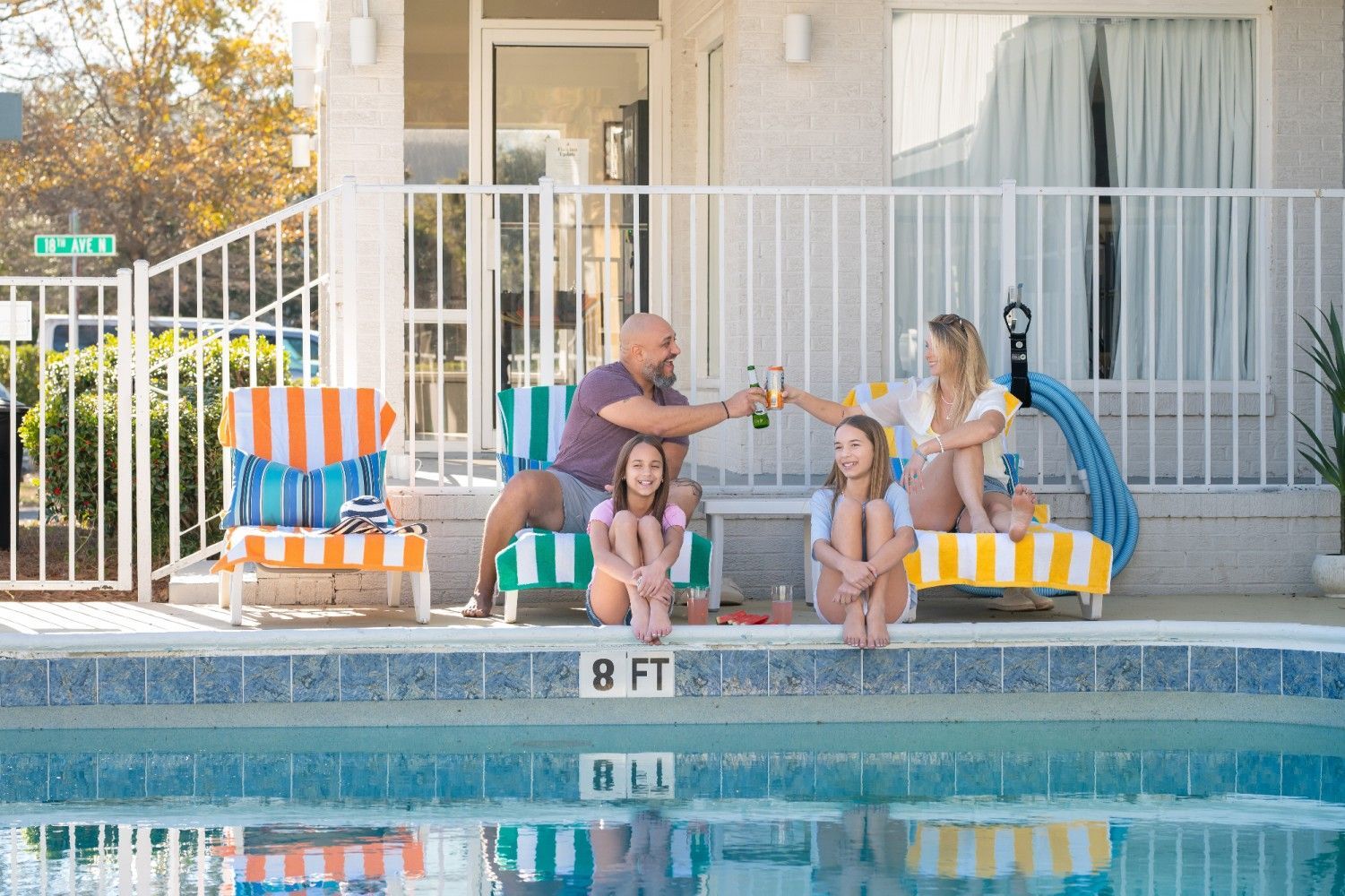 Family cheers by a pool. Adults toast beers, two girls sit, surrounded by chairs, towels. White building.