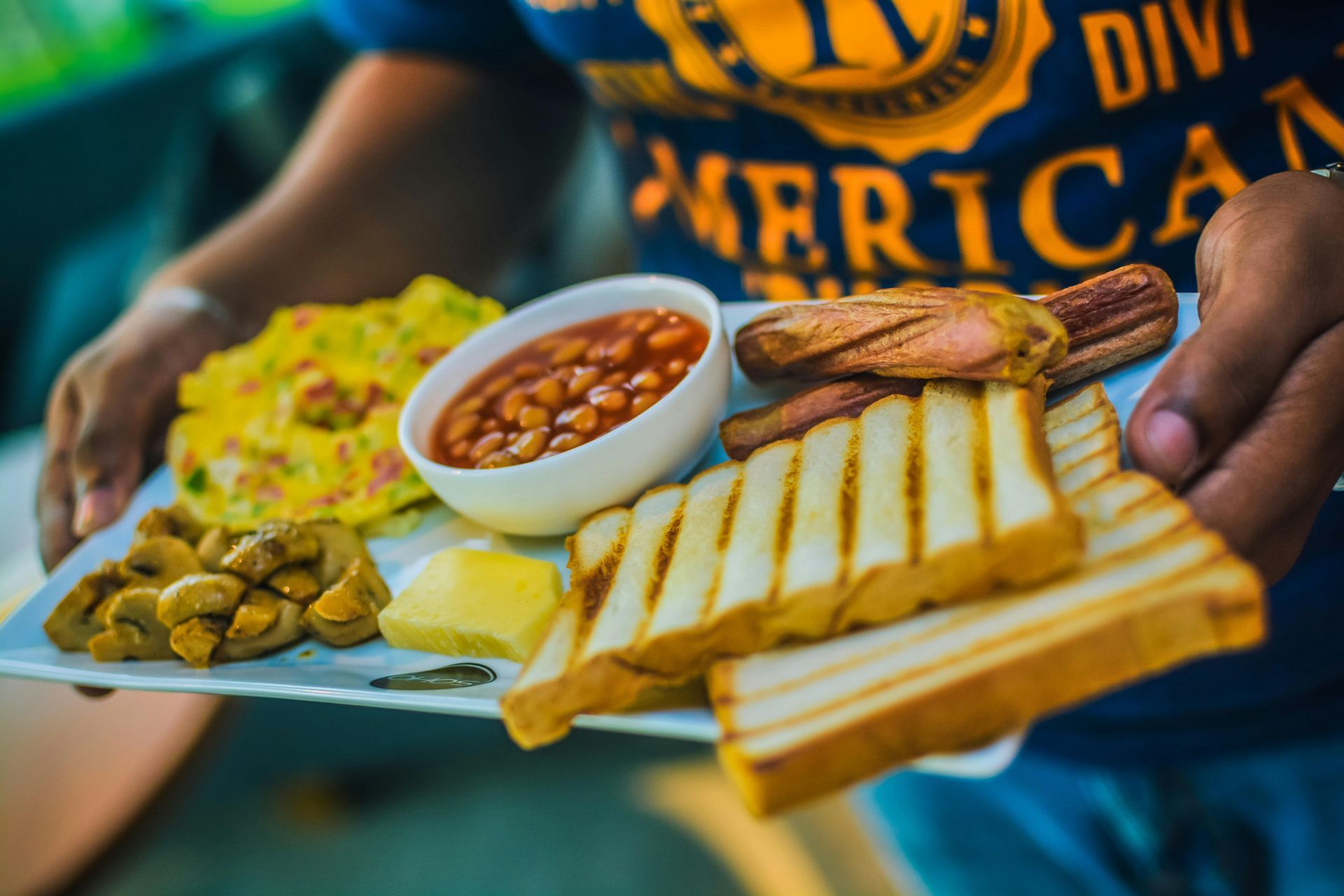 Person holding a plate of breakfast food: scrambled eggs, baked beans, toast, sausage, and mushrooms.