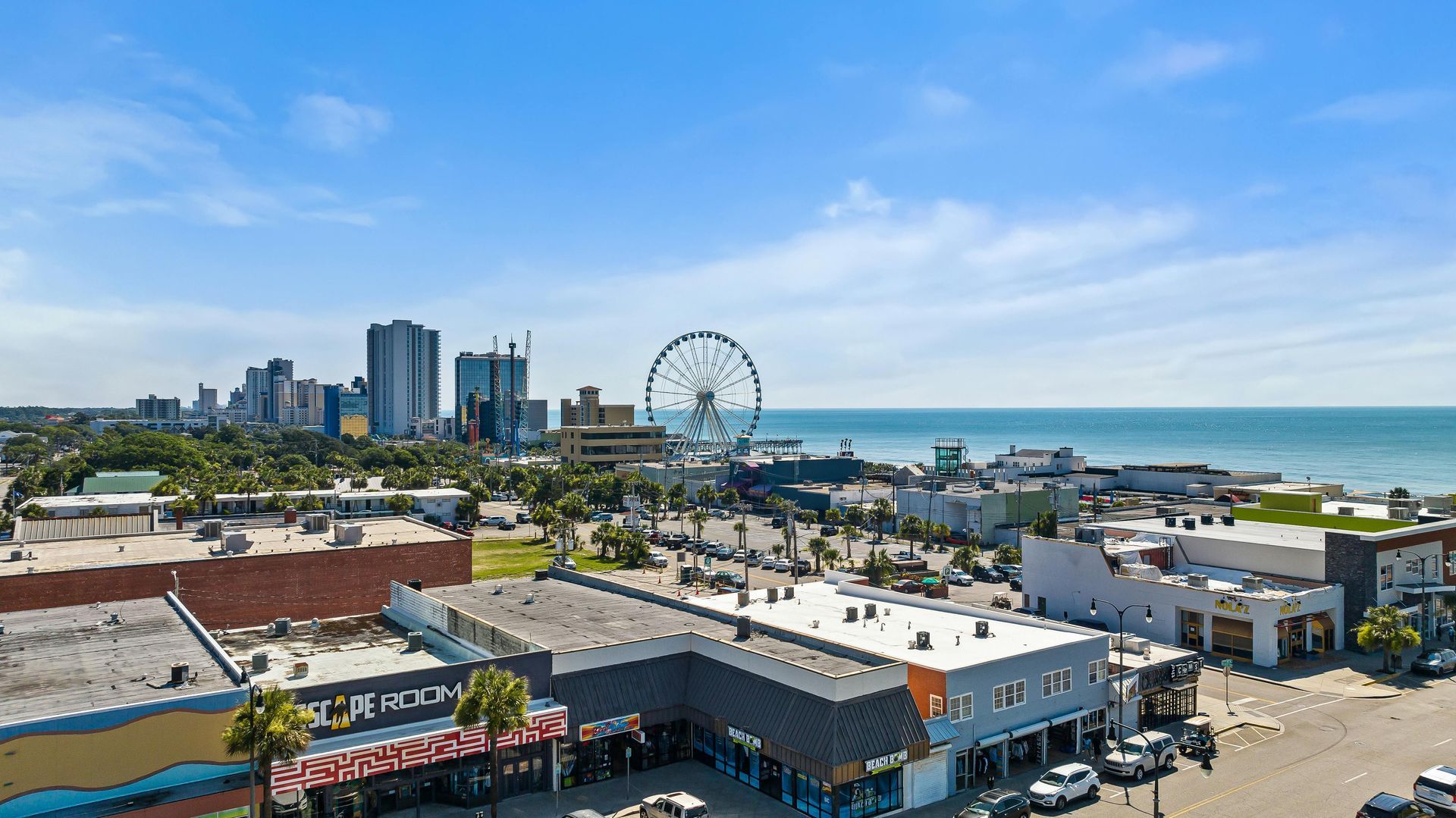 Coastal cityscape with Ferris wheel, buildings, and ocean under a blue sky.