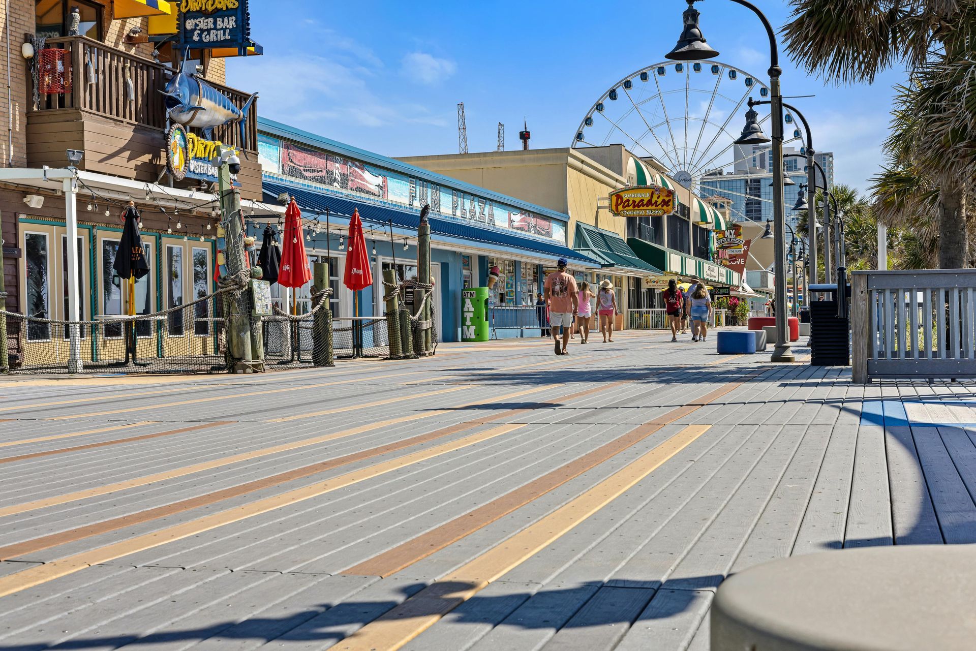 Boardwalk with shops, people strolling, Ferris wheel in background, sunny day.