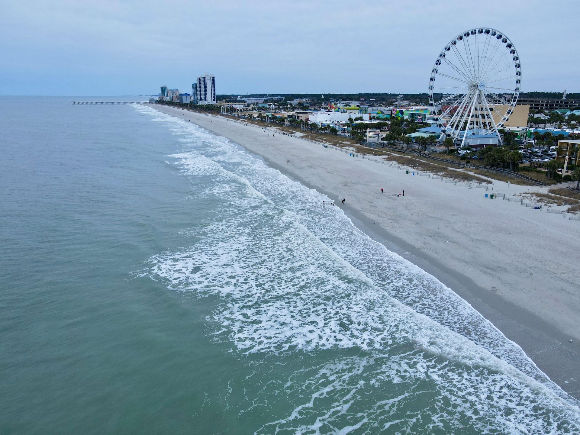 Oceanfront view of a sandy beach, buildings, and a Ferris wheel under an overcast sky.