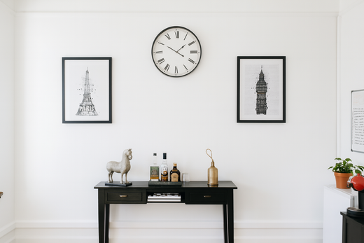 Pared blanca con una consola negra que muestra decoración, obras de arte y un reloj.