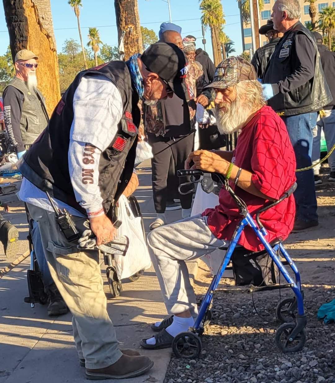 Man kneels to talk to elderly man using a walker. Men gather outdoors.