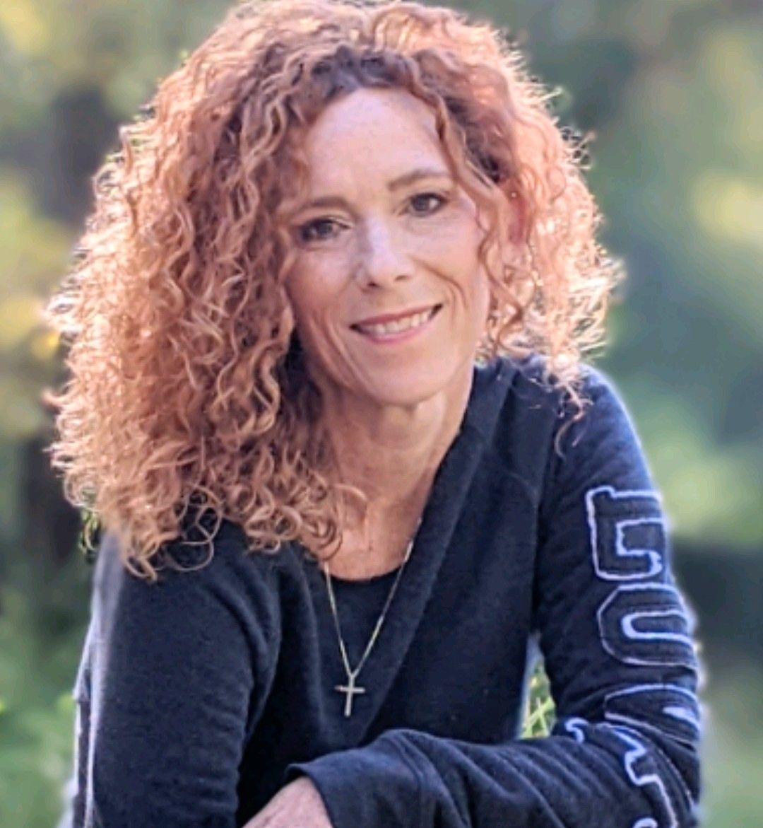 Woman with curly red hair, smiling, wearing a dark sweatshirt with the word “Gold” on the sleeve, outdoors.