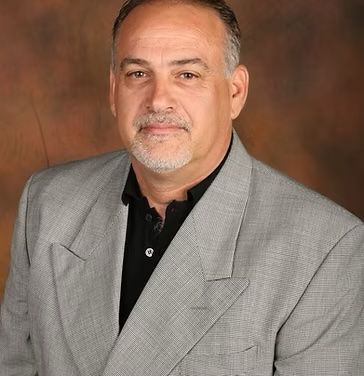 A man in a suit and black shirt is sitting in front of a brown background.