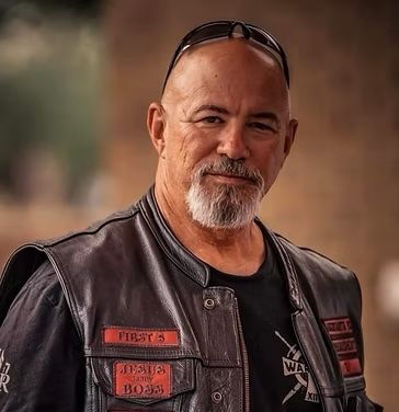 A bald man with a beard is wearing a leather vest and sunglasses.