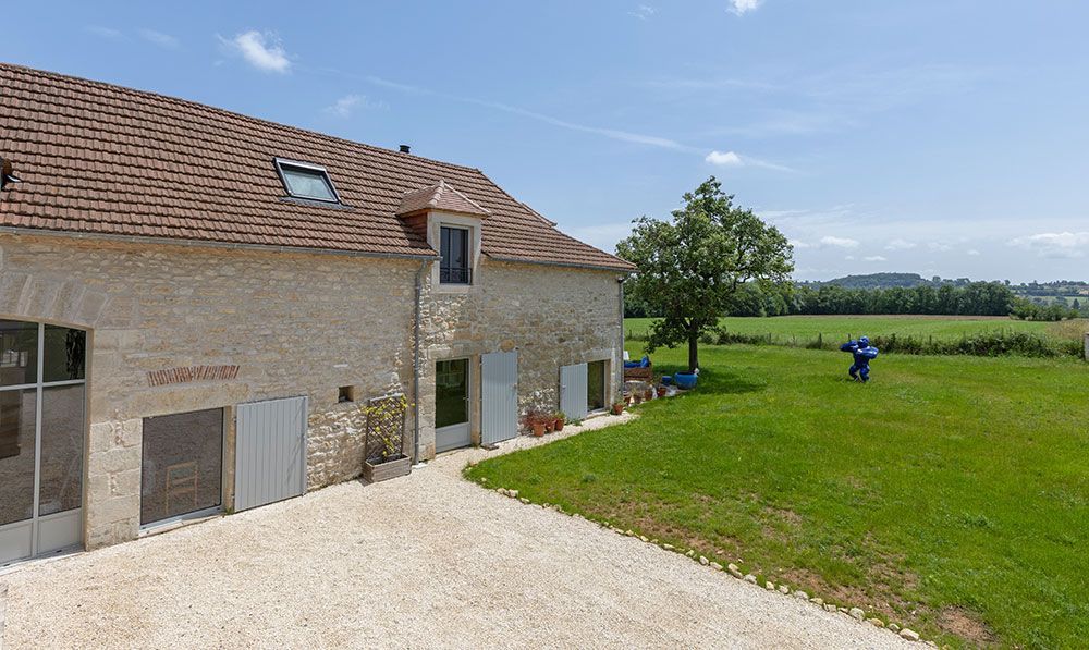 Bâtiment en pierre avec allée en gravier, pelouse verte et arbre sous un ciel bleu.