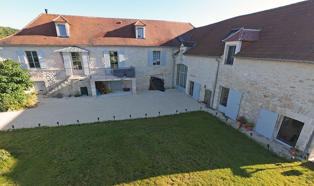 Maison en pierre avec toit rouge, volets bleu clair, et cour, pelouse gazonnée.
