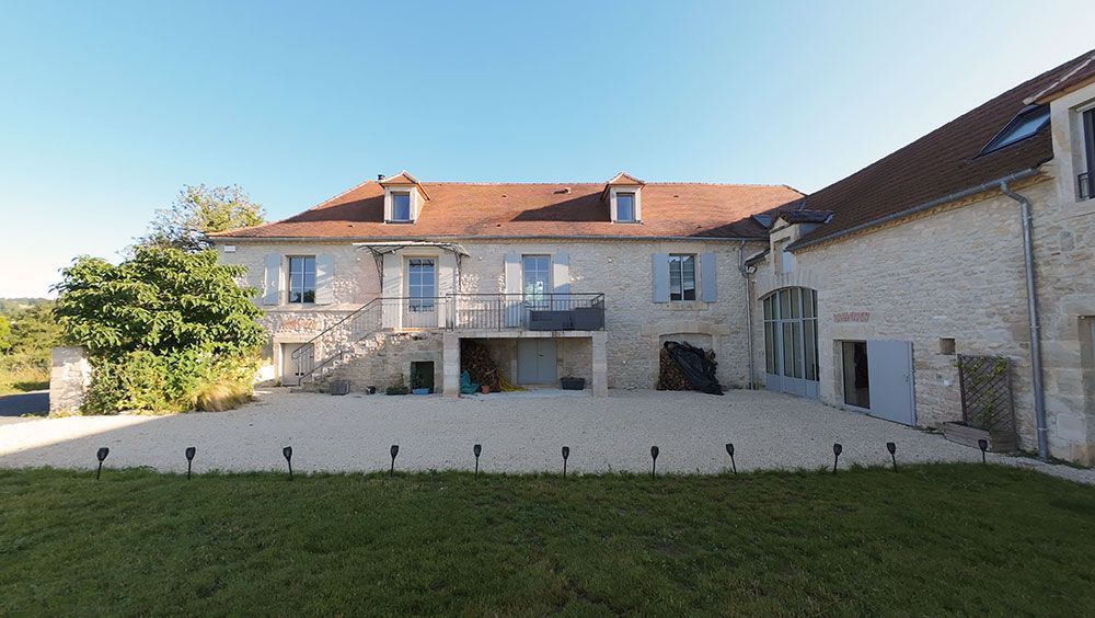 Maison en pierre avec toit de tuiles rouges et volets bleus, dressée sur un terrain herbeux sous un ciel bleu.