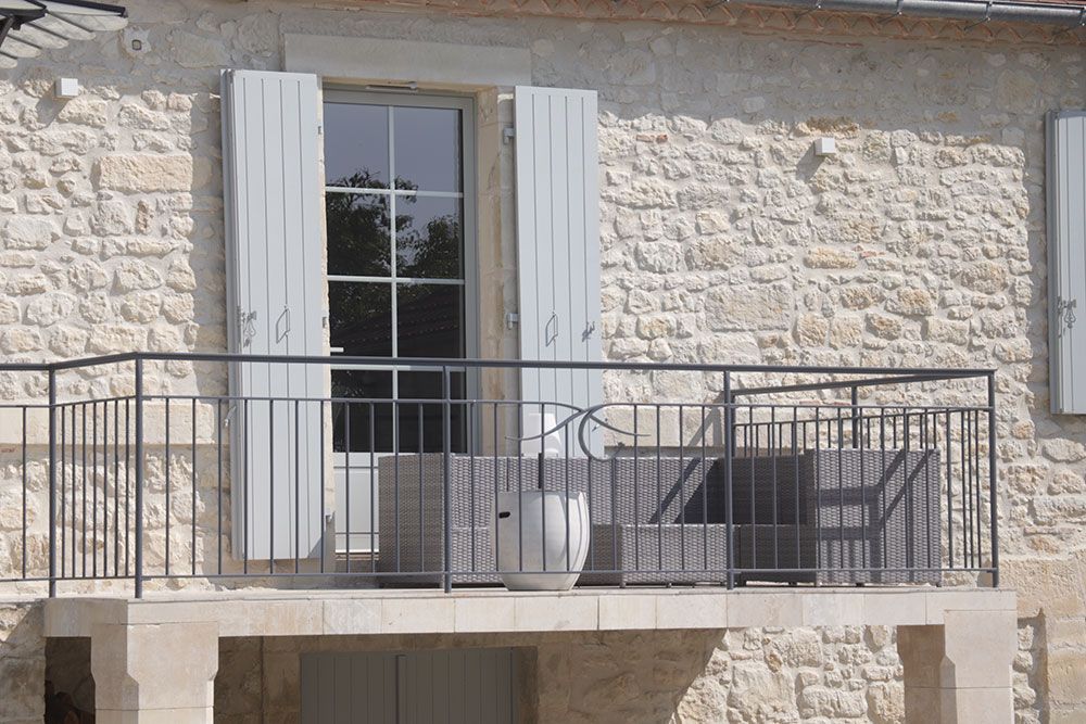 Balcon avec volets gris et garde-corps en fer forgé; extérieur du bâtiment en pierre.