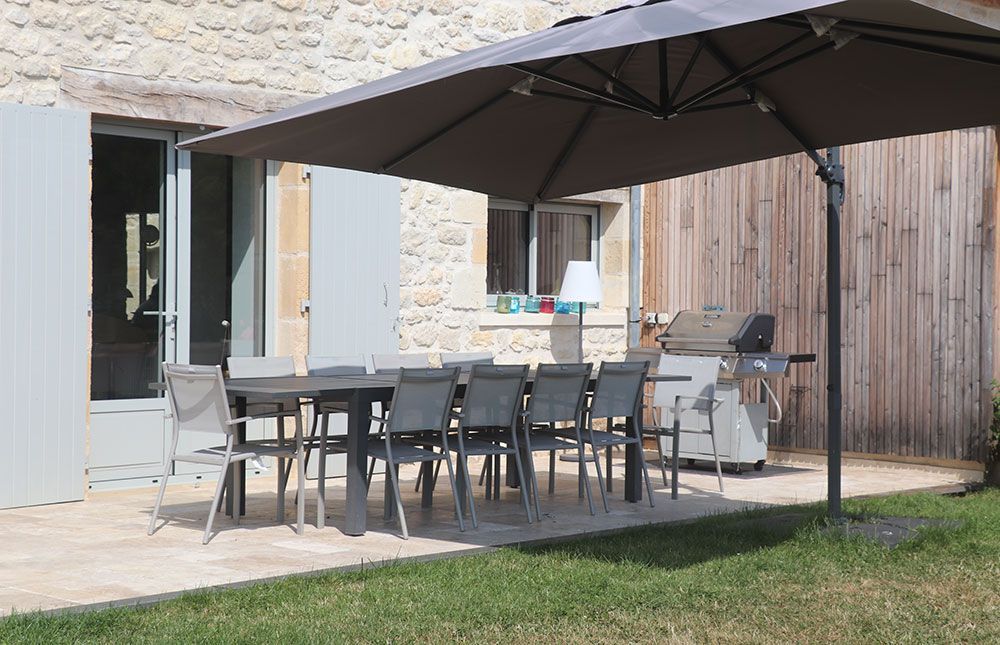 Patio avec table, chaises, parasol et barbecue près d'un bâtiment en pierre avec portes ouvertes et mur en bois.