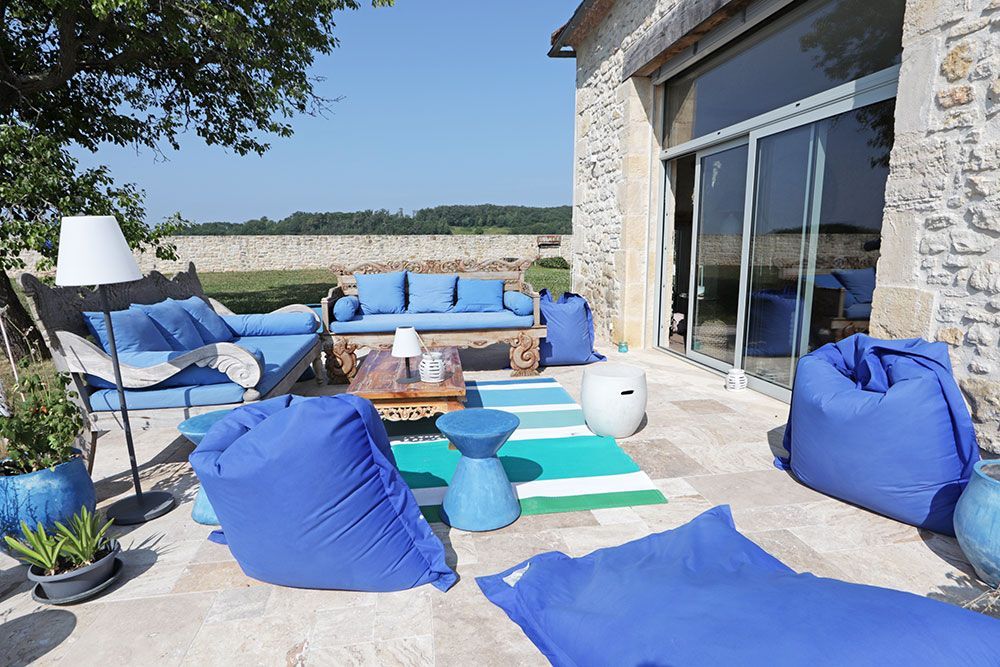 Terrasse extérieure avec mobilier bleu et poufs près d'un bâtiment en pierre ; journée ensoleillée.