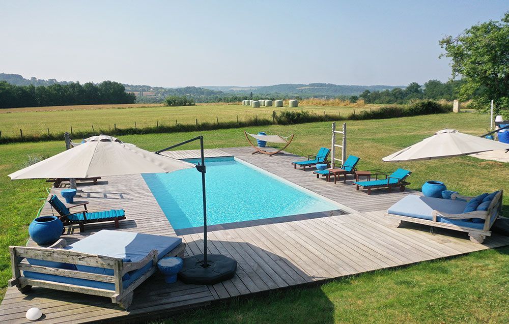 Piscine avec chaises longues et parasols sur une terrasse en bois, avec vue sur un champ et des collines au loin.