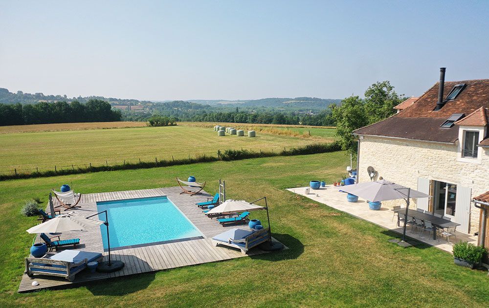 Une piscine et un patio à côté d'une maison en pierre, donnant sur un champ herbeux.