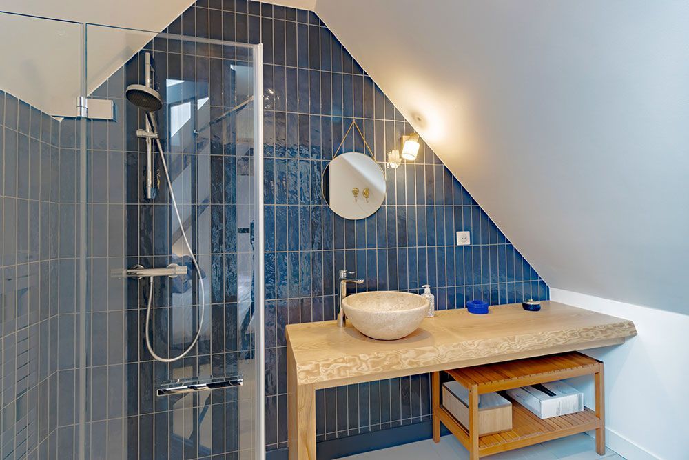 Salle de bain avec murs carrelés bleus, douche, lavabo sur un plan de travail en bois sous un plafond en pente.