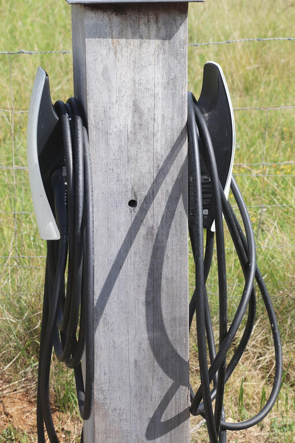 Deux câbles de recharge pour véhicules électriques sont suspendus à un poteau en bois dans un cadre extérieur herbeux.