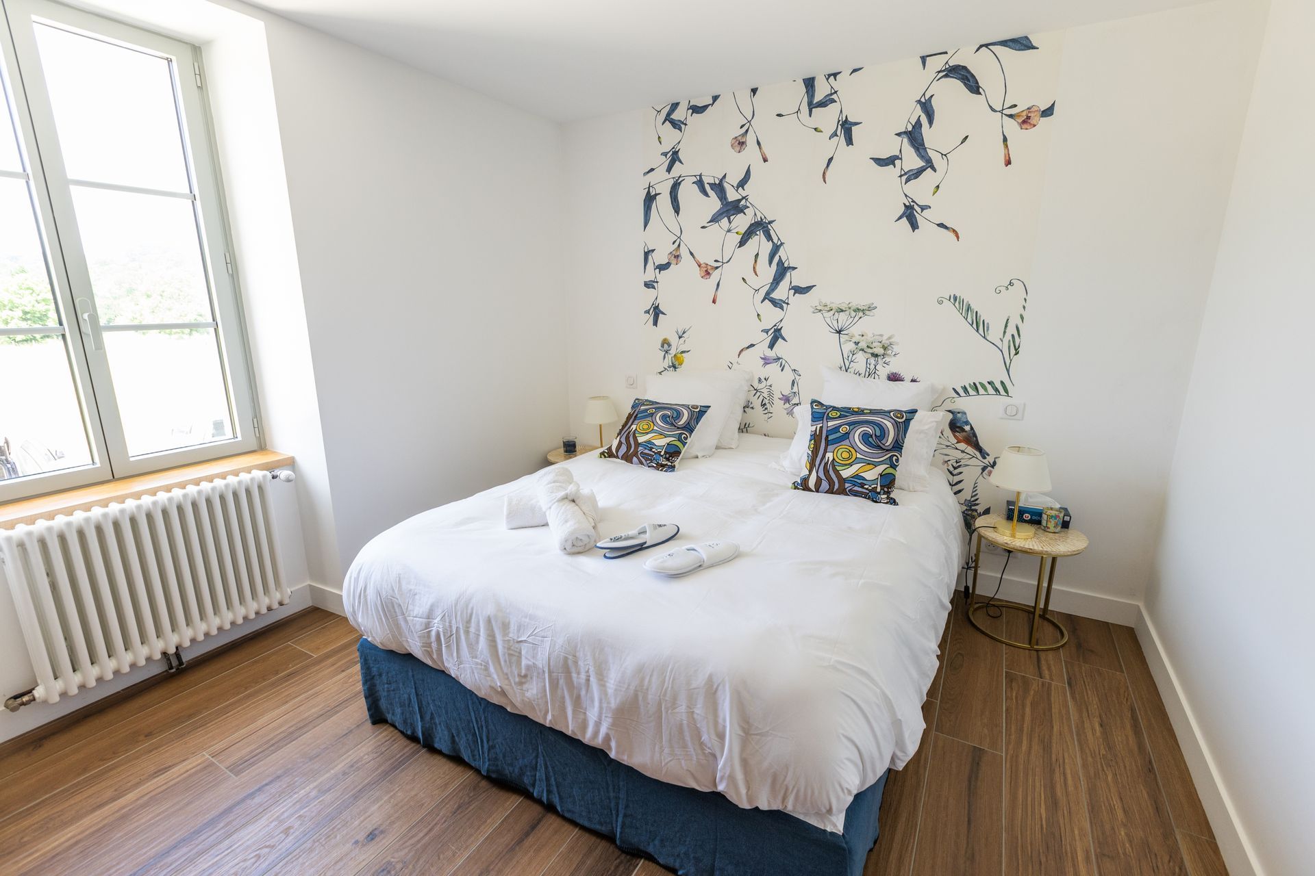 Chambre avec literie blanche, jupe de lit bleue, mur d'accent floral, plancher de bois, fenêtre.