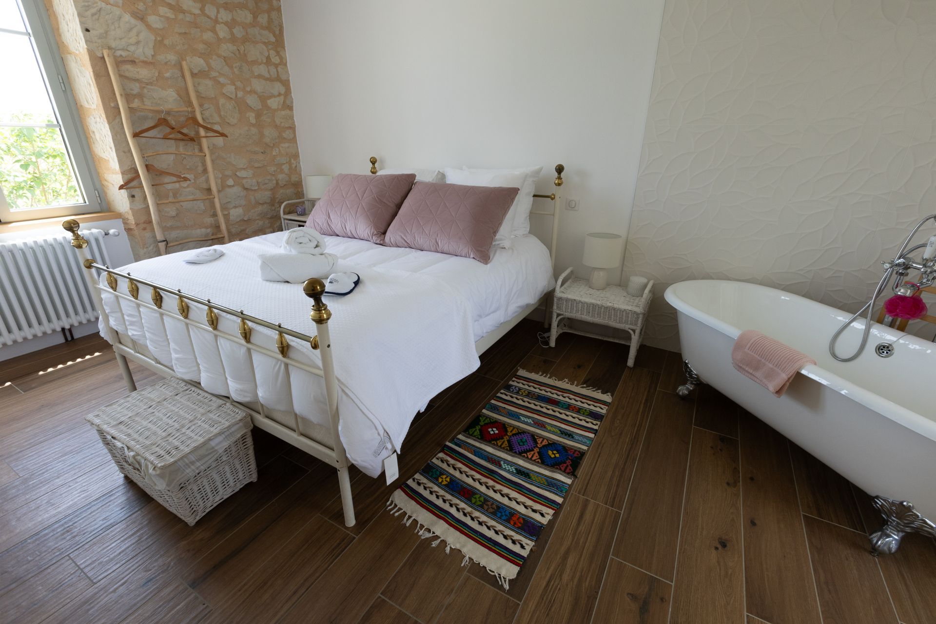 Une chambre lumineuse avec un lit blanc, des oreillers roses et une baignoire sur pieds. Un petit tapis recouvre le parquet.