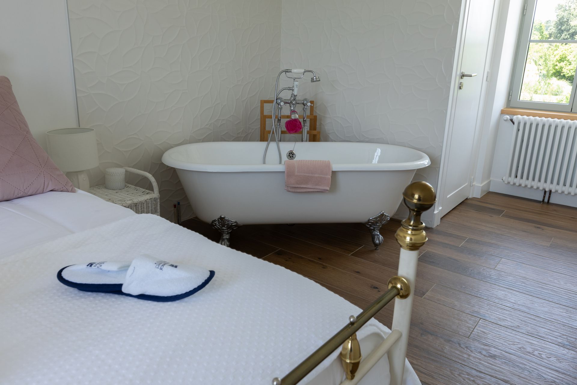 Chambre avec baignoire sur pieds, lit avec oreiller rose, pantoufles blanches et fenêtre avec vue.