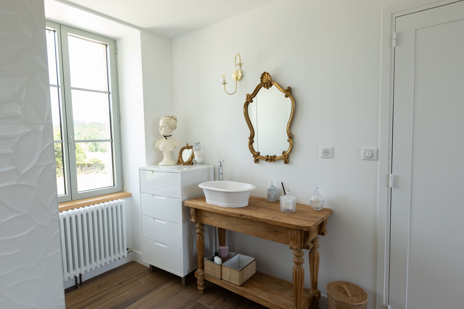 Salle de bain avec murs blancs, une console en bois avec un lavabo, un miroir à cadre doré et une fenêtre.