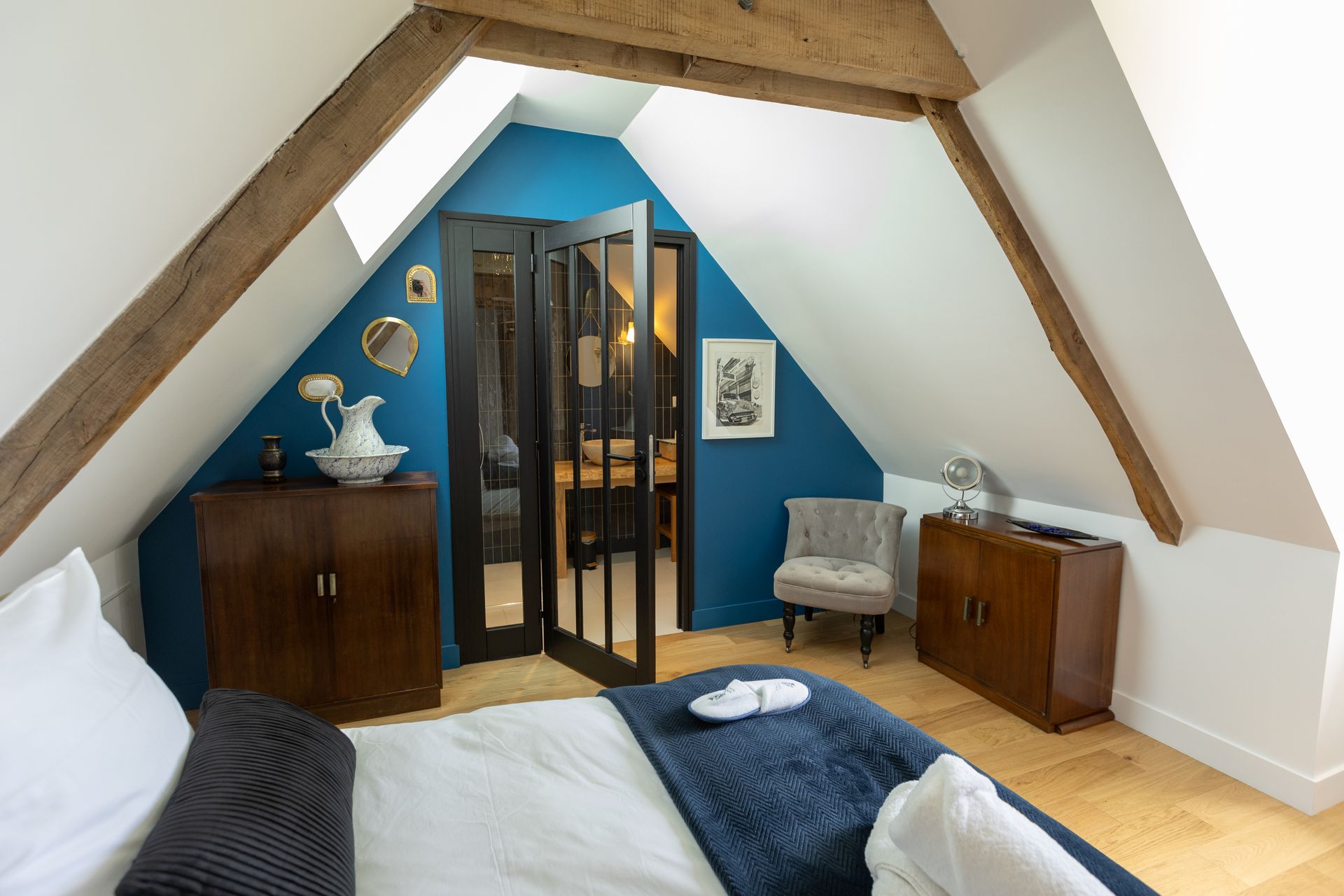Chambre dans un grenier avec mur d'accent bleu, poutres en bois, lit et portes-fenêtres ouvertes menant à une salle de bain.