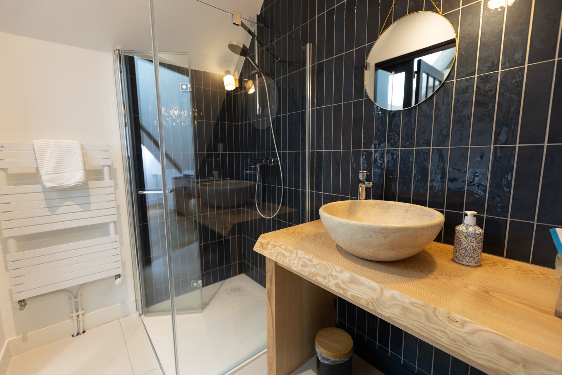 Salle de bain moderne avec murs carrelés bleu foncé, lavabo en pierre sur une vanité en bois et douche en verre.