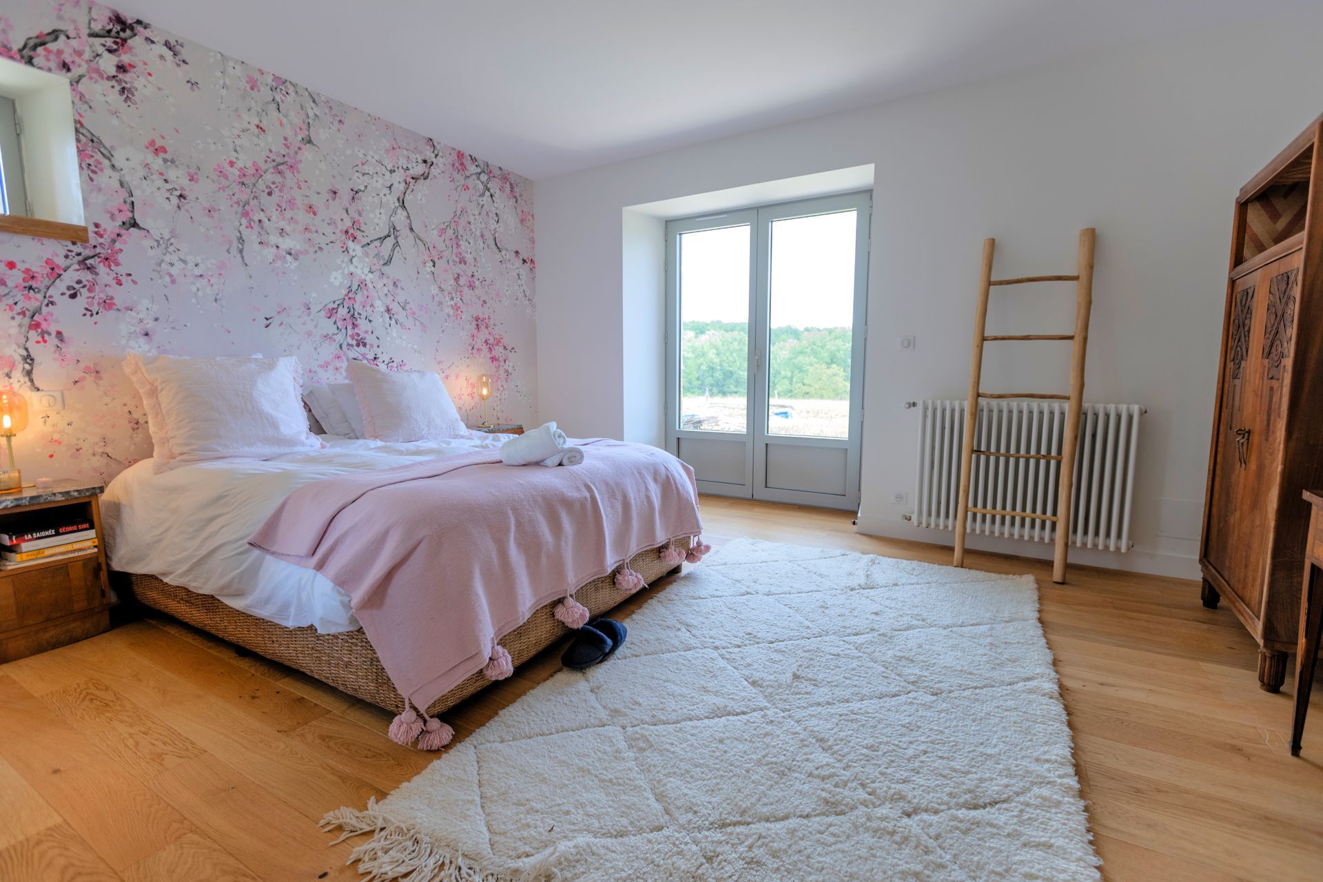 Chambre avec papier peint fleuri, lit avec couverture rose, tapis, parquet, fenêtre et meubles en bois.