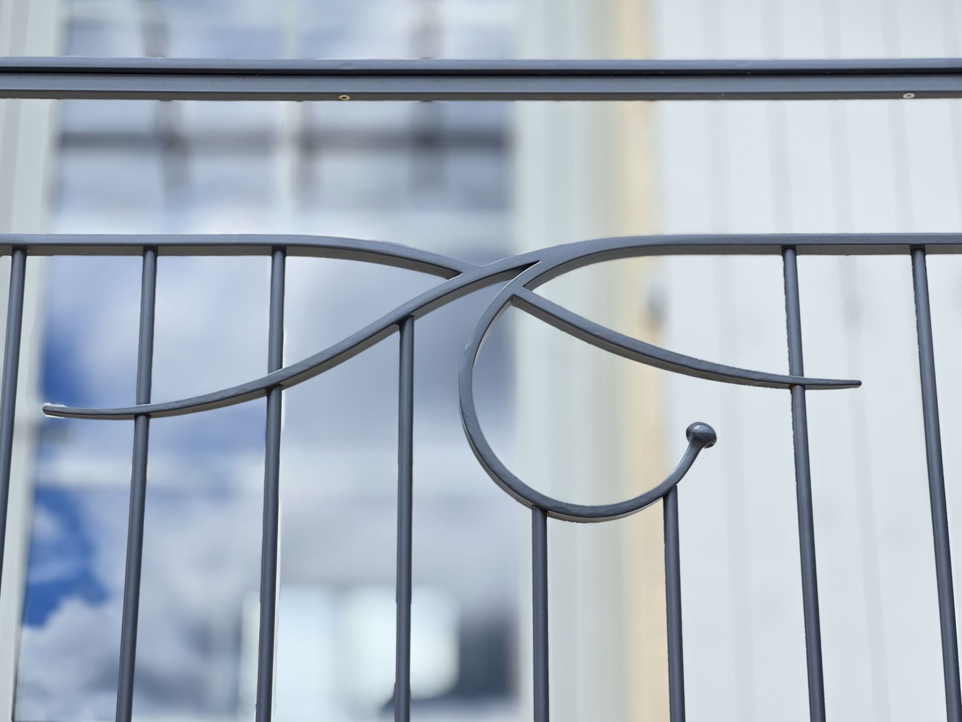 Gros plan d'une balustrade en métal gris ornée d'un motif décoratif en volutes. L'arrière-plan montre un bâtiment blanc flou.