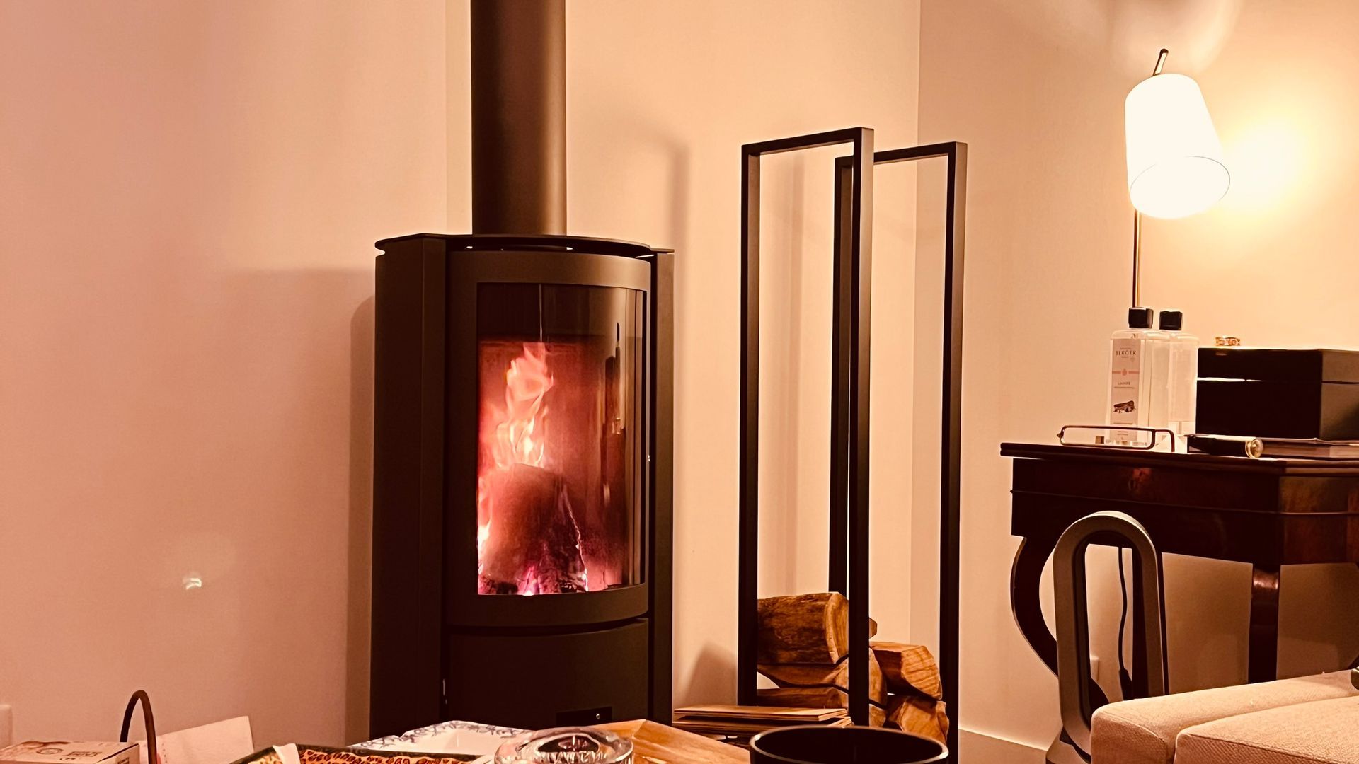 Cheminée avec des flammes brûlantes dans une pièce avec des miroirs décoratifs, des bûches et un bureau avec une lampe.