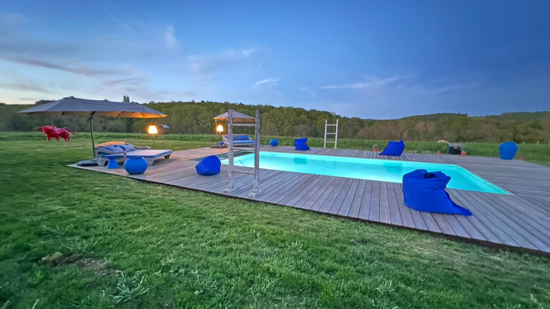 Piscine avec terrasse en bois et sièges bleus sur un terrain herbeux au crépuscule.