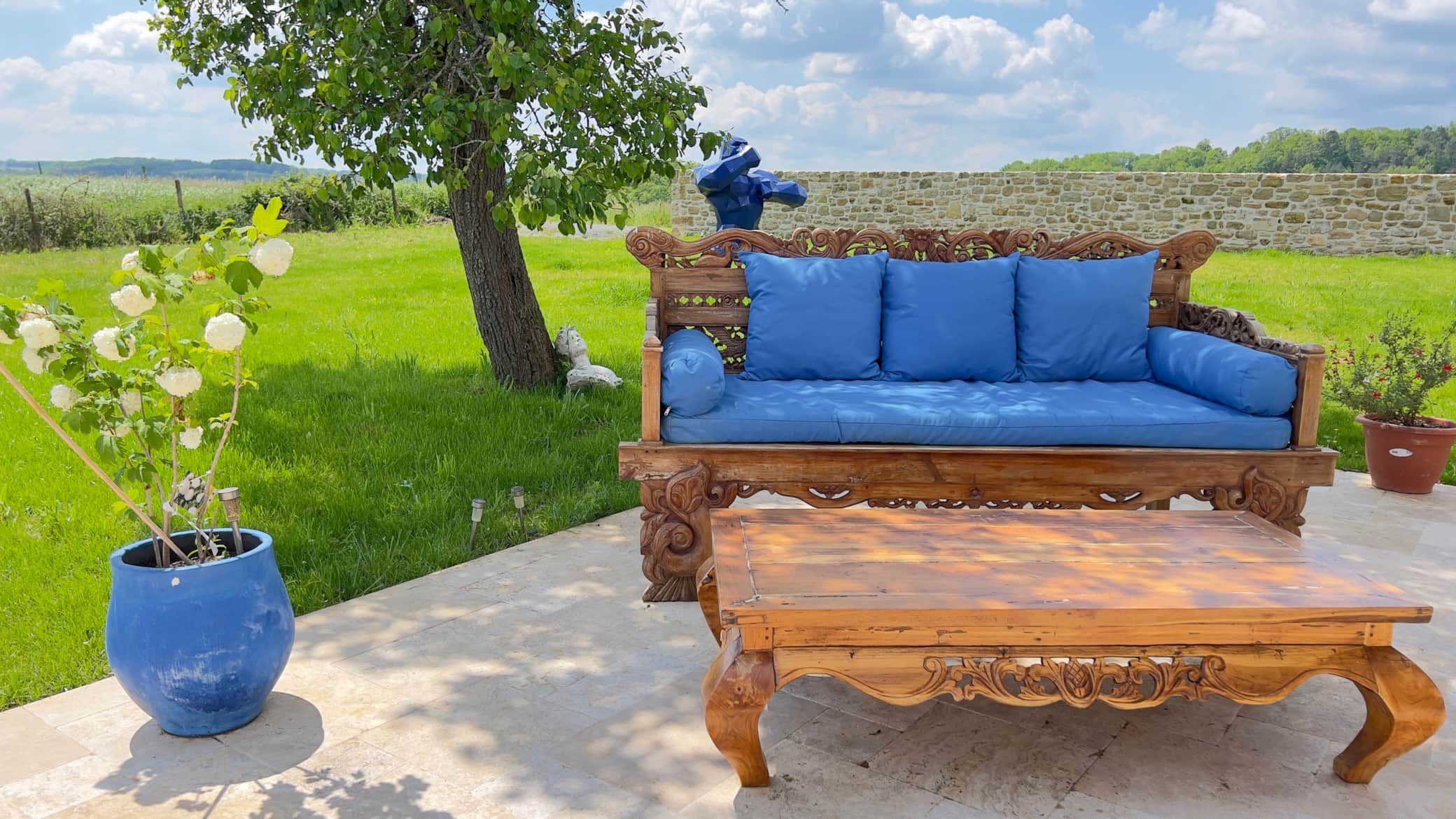 Sièges d'extérieur en bois avec coussins bleus et table basse assortie sur une terrasse donnant sur un champ herbeux.