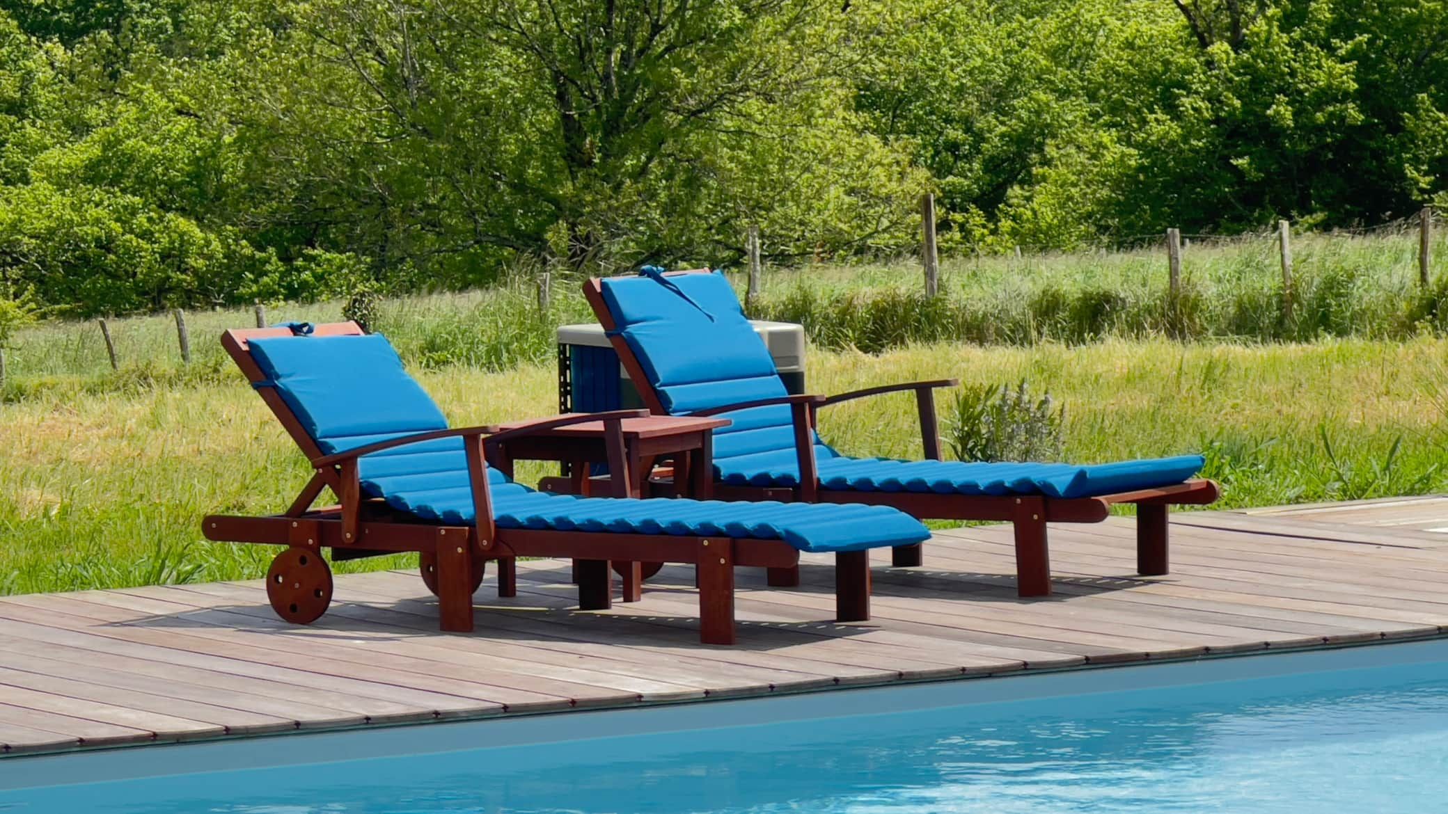 Deux chaises longues en bois avec des coussins bleus à côté d'une piscine, avec une zone boisée en arrière-plan.