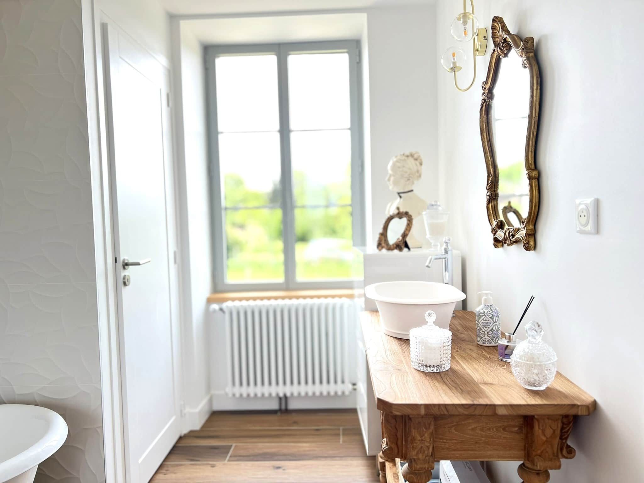 Salle de bain avec vanité en bois, miroir doré, fenêtre donnant sur un champ vert et luminaires blancs.