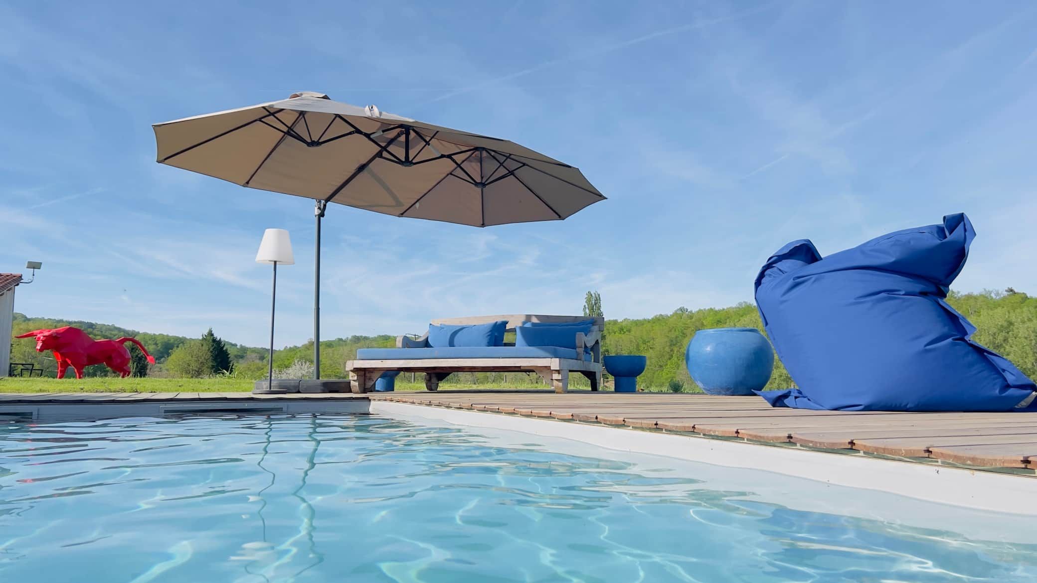 Scène au bord de la piscine avec des sièges bleus, un parasol et une sculpture de taureau rouge sous un ciel bleu vif.