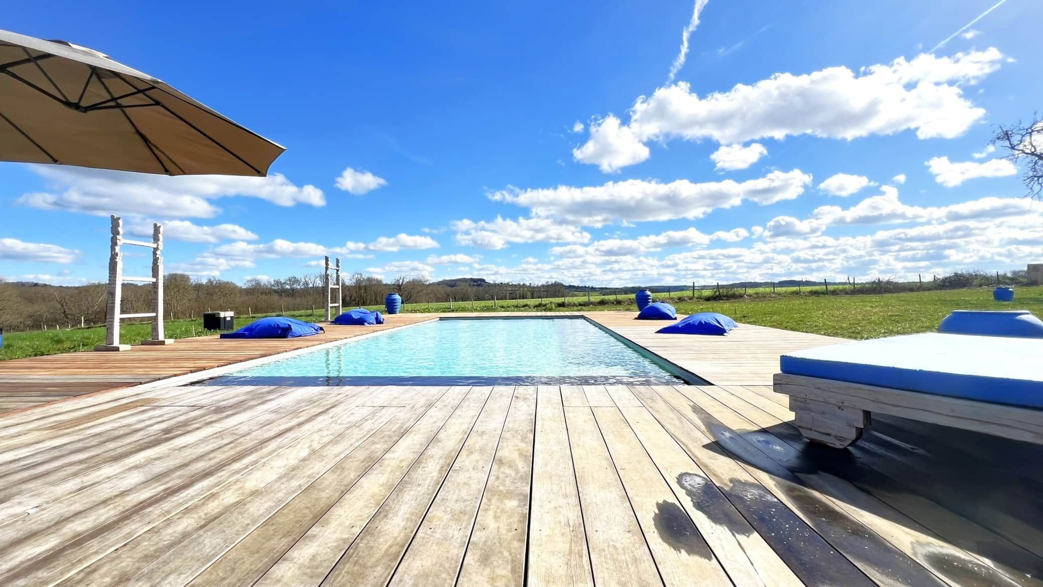 Terrasse en bois avec une piscine rectangulaire, des poufs bleus et un ciel ensoleillé et partiellement nuageux.