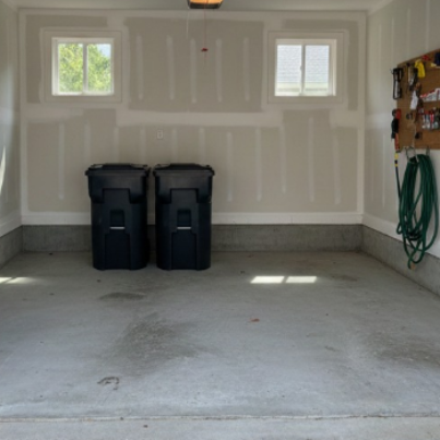 Empty garage with two black trash cans, two windows, and a hose on the wall.
