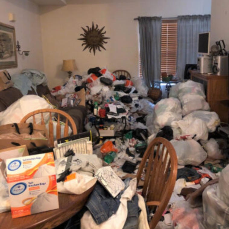 Cluttered living room. Piles of clothes and bags cover furniture, obstructing view. Dark wooden table, light walls.