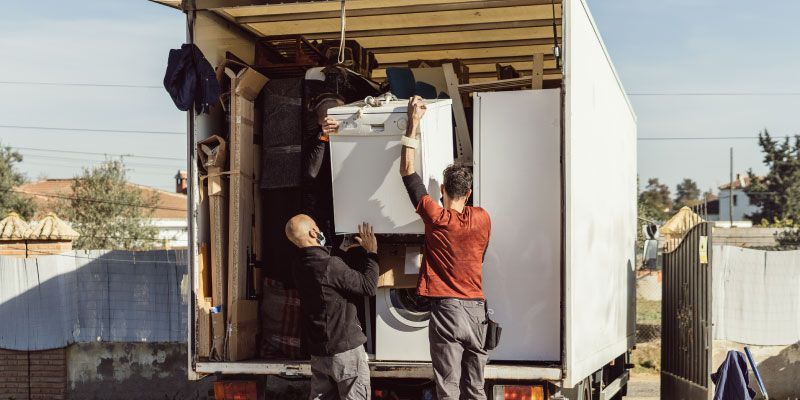 Two people load a refrigerator into a moving truck. The truck is white, parked outdoors.