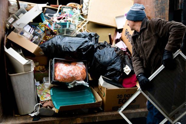 Person loading trash into a dumpster, holding a window screen. Dumpster overflowing with garbage.