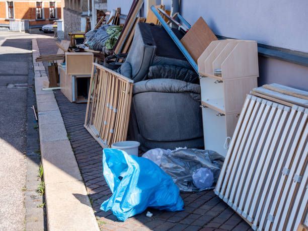 Pile of discarded furniture and debris on a sidewalk next to a road; blue trash bag in front.