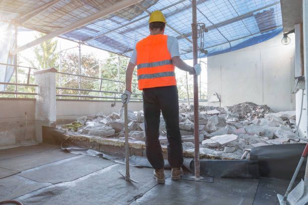 Construction worker in orange vest and hard hat surveying a rubble-filled interior, holding tools.