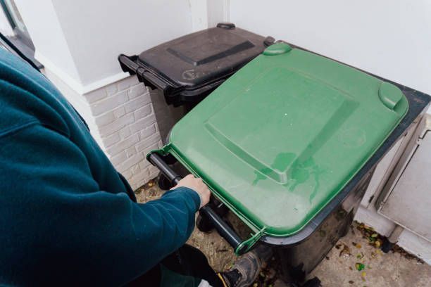 Person lifting the lid of a green trash bin, another black bin in the background, by a white wall.