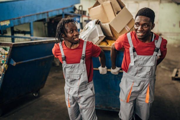 Two people in red shirts and gray overalls lifting a large blue bin filled with cardboard boxes.