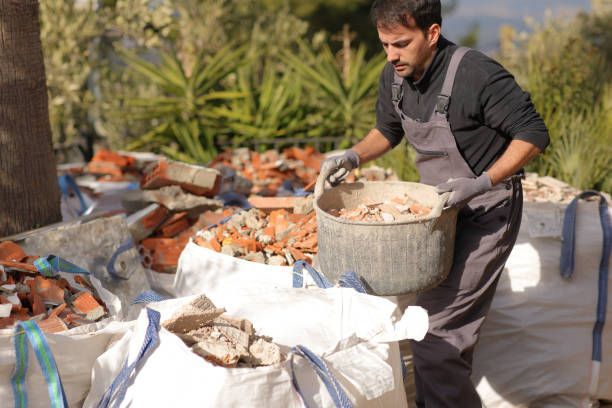 Man in overalls and gloves filling a bin with rubble, putting it into white bags outside.