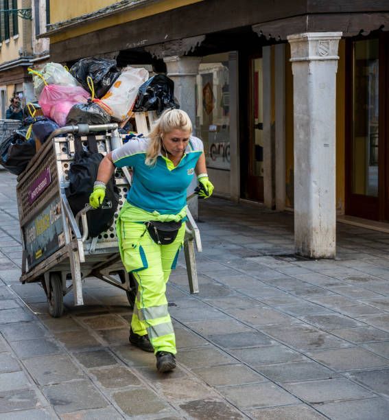 Woman in neon workwear pulling a trash cart down a cobblestone street.