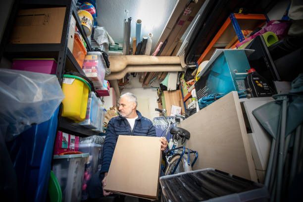 Man in cluttered storage room holding cardboard. Shelves filled with boxes, bins, and items.