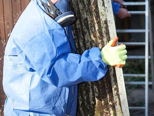 Person in blue protective suit and respirator, carrying asbestos roofing panel.