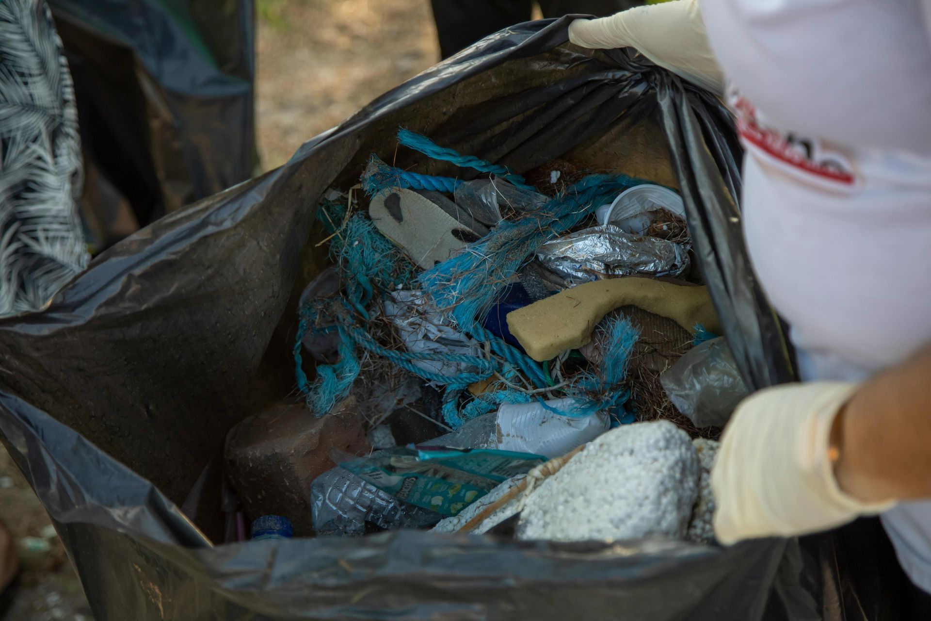 Person wearing gloves holding a black trash bag filled with litter collected outdoors.